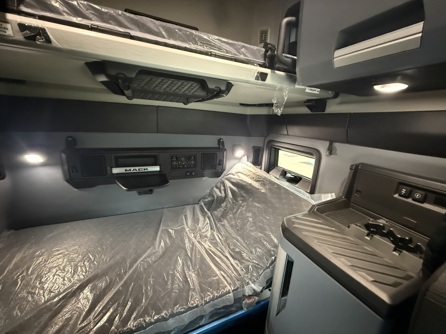 Truck cab interior showing sleeper berth area with overhead storage, control panel, and plastic-covered bed; Mack branding on dash area.