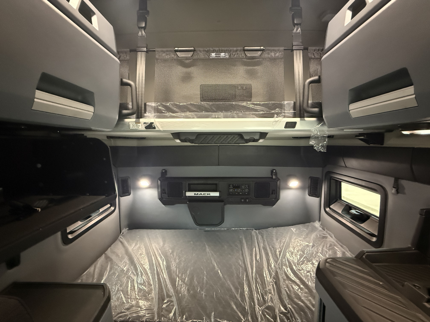 Truck cabin interior showing cargo area with sleeper bunk and control panel labeled 'MACK' in center; plastic-covered floor and seats; overhead compartments.