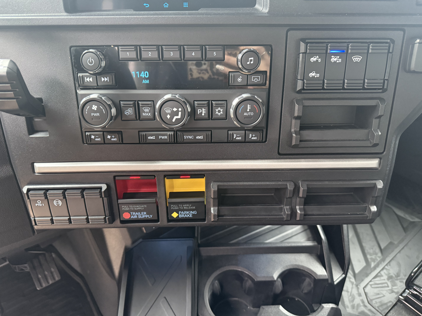 Dashboard panel of a Mack truck showing various controls, radio display 1140 AM, climate and auxiliary switches, and two colored hazard/engineering switches.