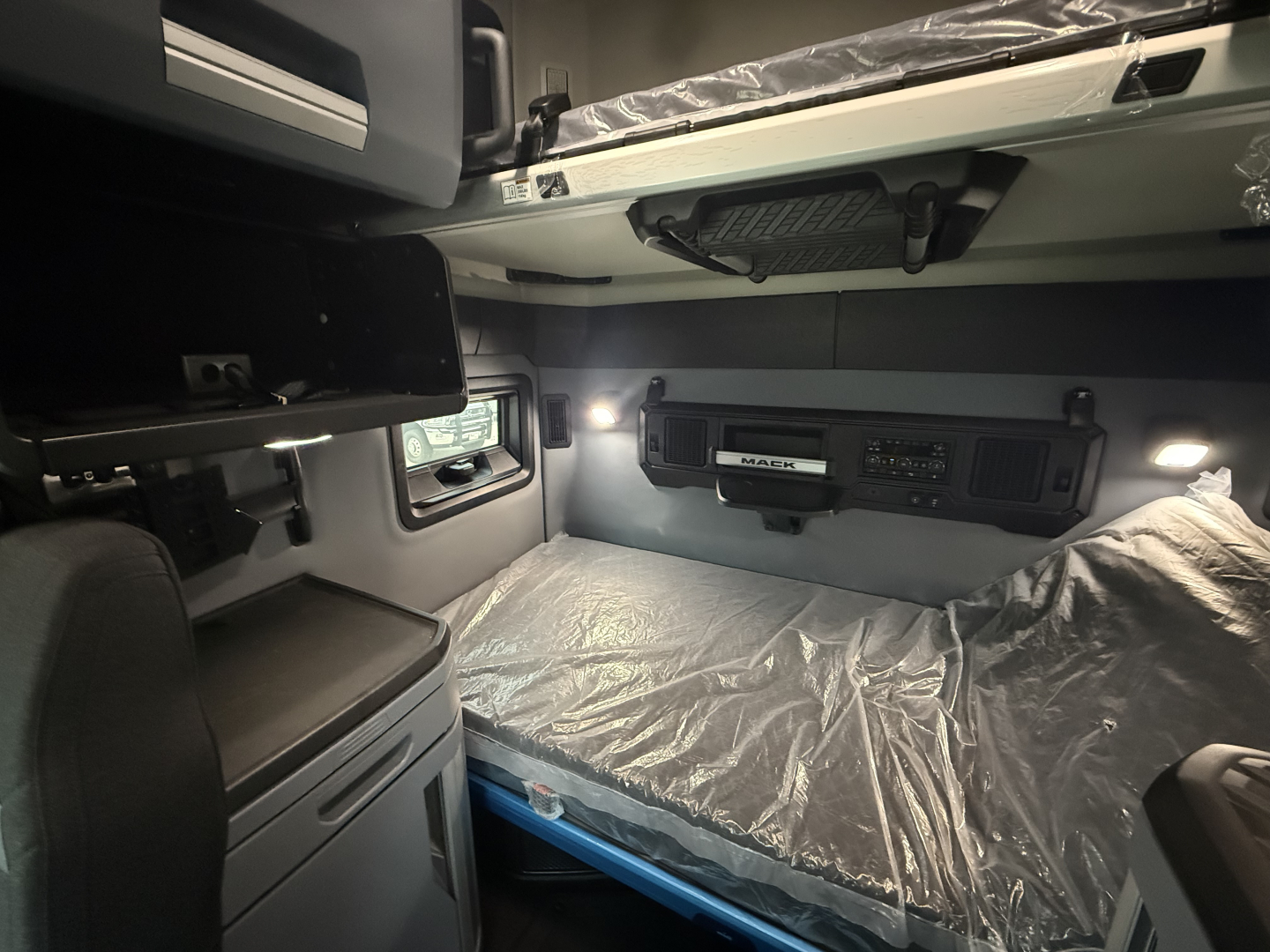 Mack PR64T76H truck interior bunk area with side cabinets and overhead storage, sleeper cab with plastic-covered bed and dim lighting.