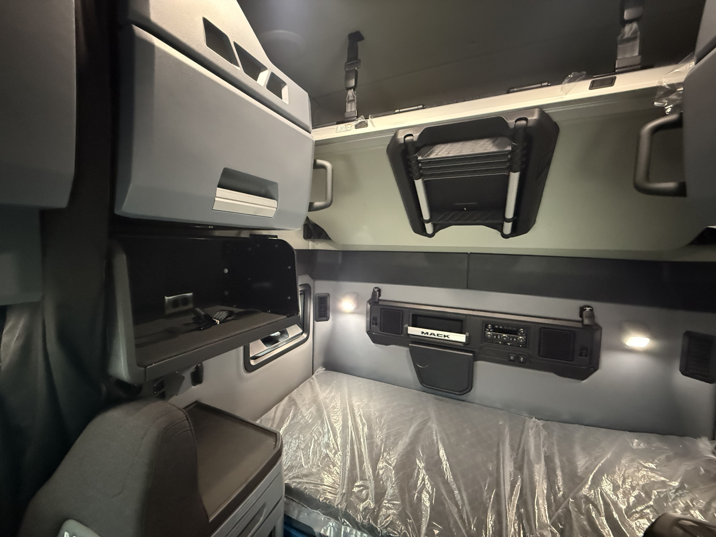 Interior sleeper cab of a Mack PR64T76H showing overhead storage pouch, wall-mounted controls, seating area, and plastic-wrapped bed surface.