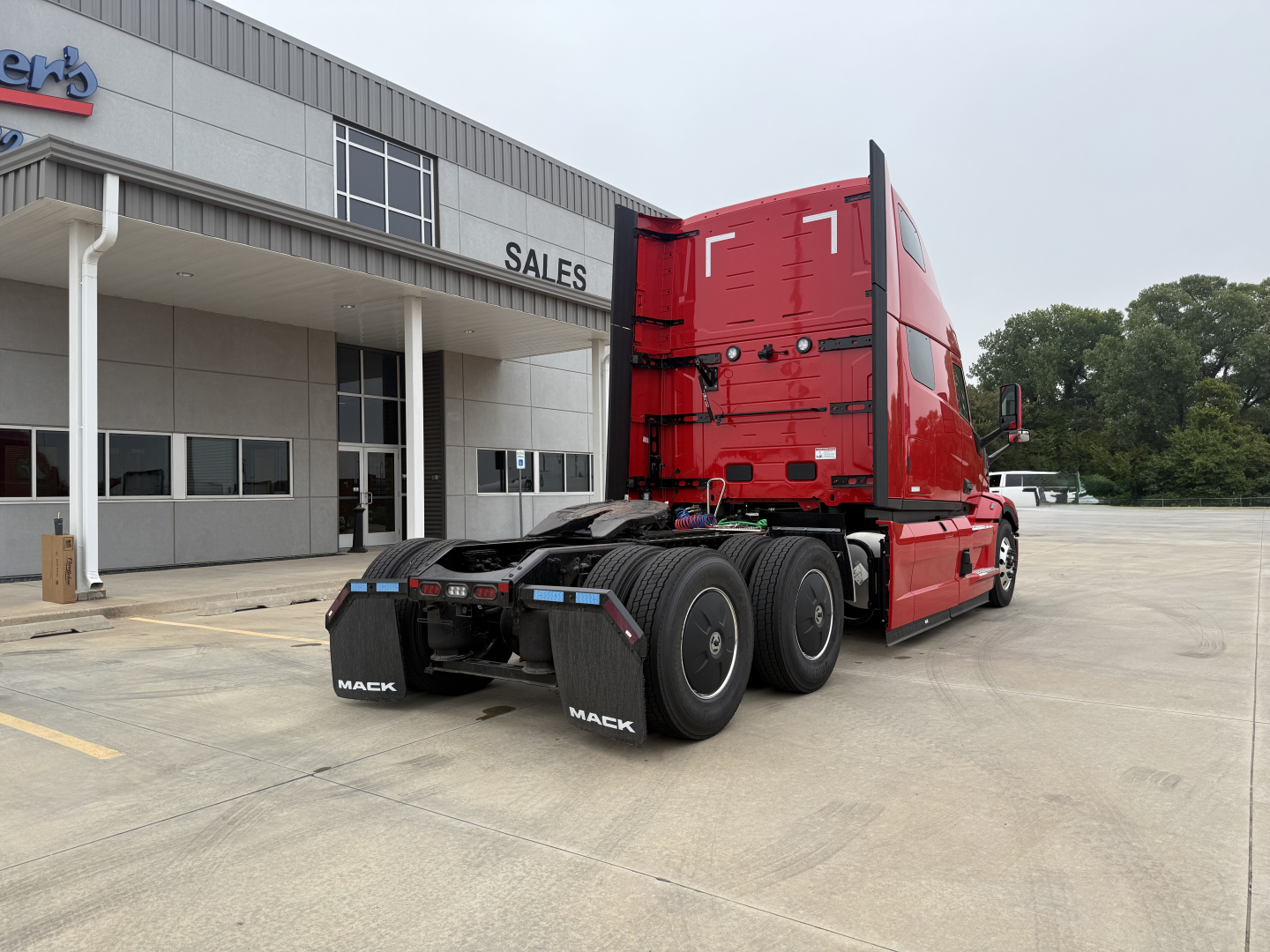 Truck tractor rear three-quarter view of a red 2026 Mack PR64T76H showing the sleeper cab, dual rear axles, and fifth-wheel area in front of a dealership showroom.