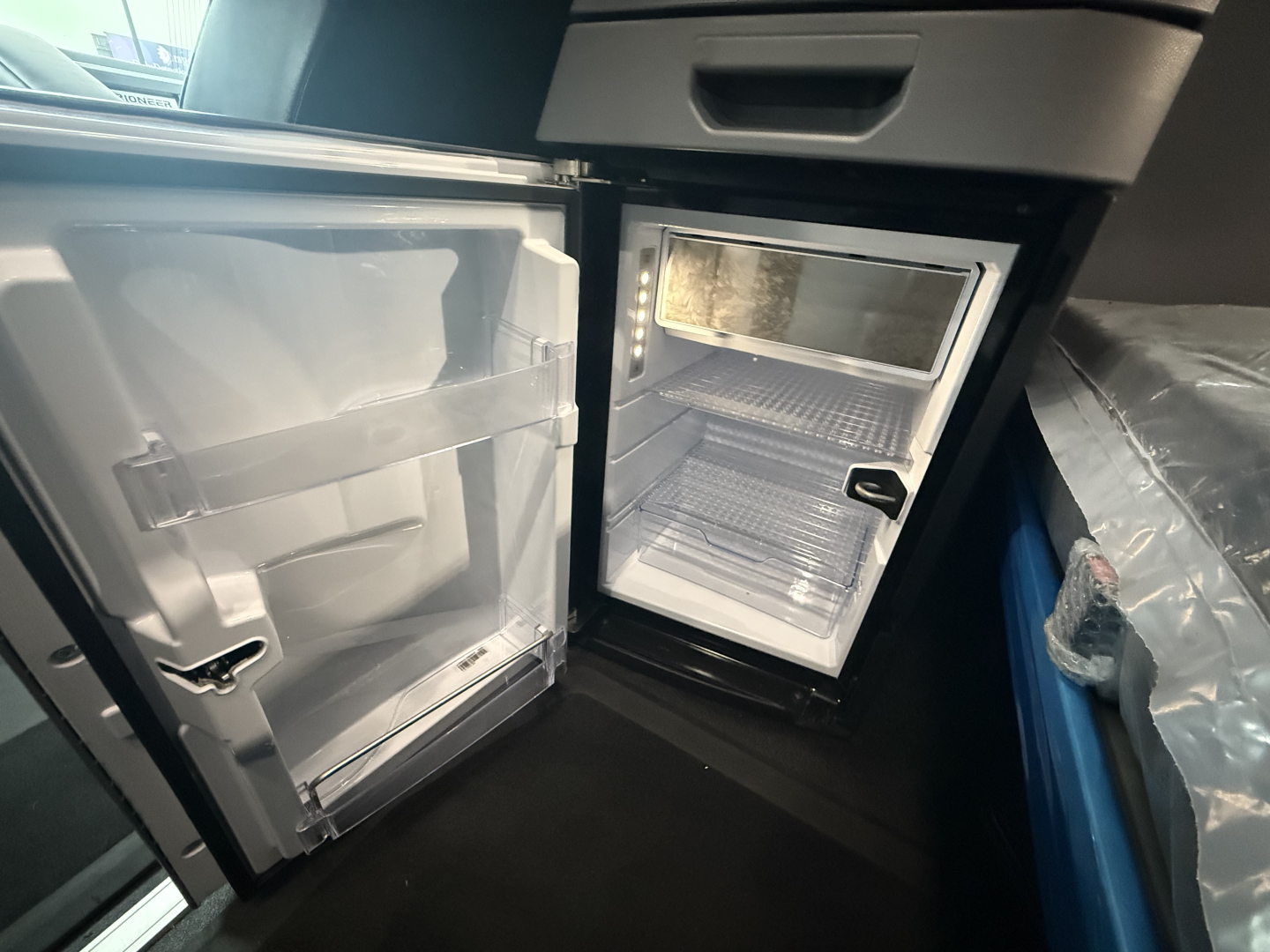 Truck cab interior showing a compact refrigerator/freezer unit with open doors and empty shelves.