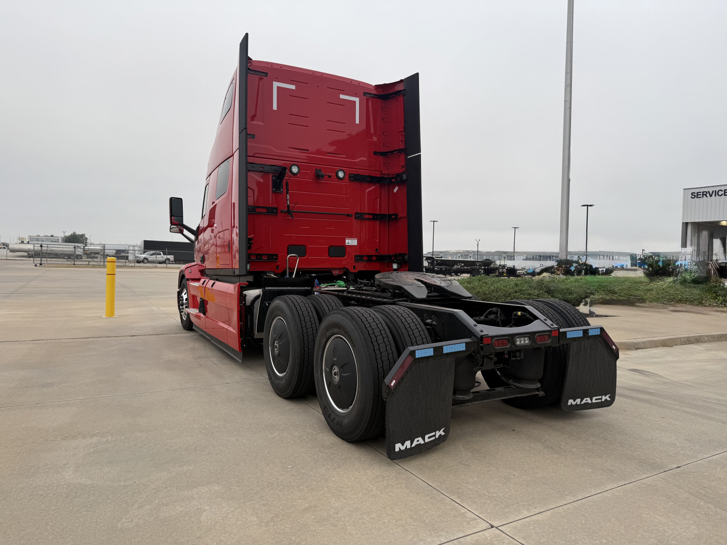 Mack PR64T76H truck in a parking lot, rear view showing three rear axles, red cab with tall sleeper, black mud flaps labeled 'MACK', fifth wheel area, no visible text on side panels.