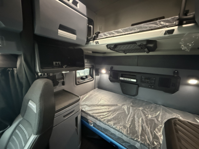 Truck cab interior of a new 2026 Mack PR64T76H showing sleeper bunk area and overhead storage with plastic wrap on bed.