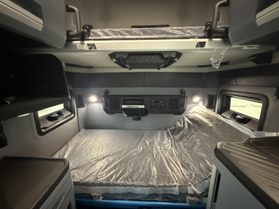 Interior cab of a new Mack PR64T76H truck showing bunk bed with plastic cover, overhead storage cabinets, and control panel on the wall.