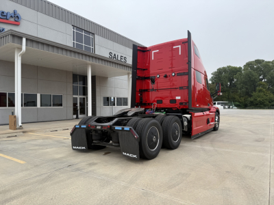 Truck tractor rear three-quarter view of a red 2026 Mack PR64T76H showing the sleeper cab, dual rear axles, and fifth-wheel area in front of a dealership showroom.