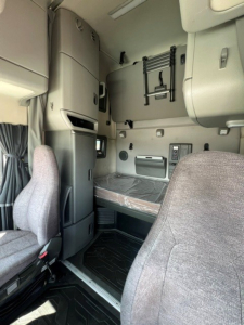 Volvo VNL64T760 sleeper cab interior showing bunk bed area and control panel. Includes passenger seat in foreground, overhead storage, and ladder/steps near window.