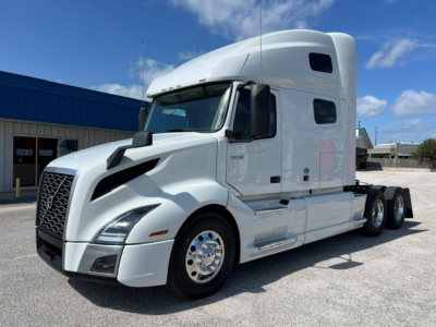 Truck cab showing Volvo VNL64T760 sleeper with white paint, aerodynamic hood, dual rear axles, and chrome wheels; side mirrors and window details visible.