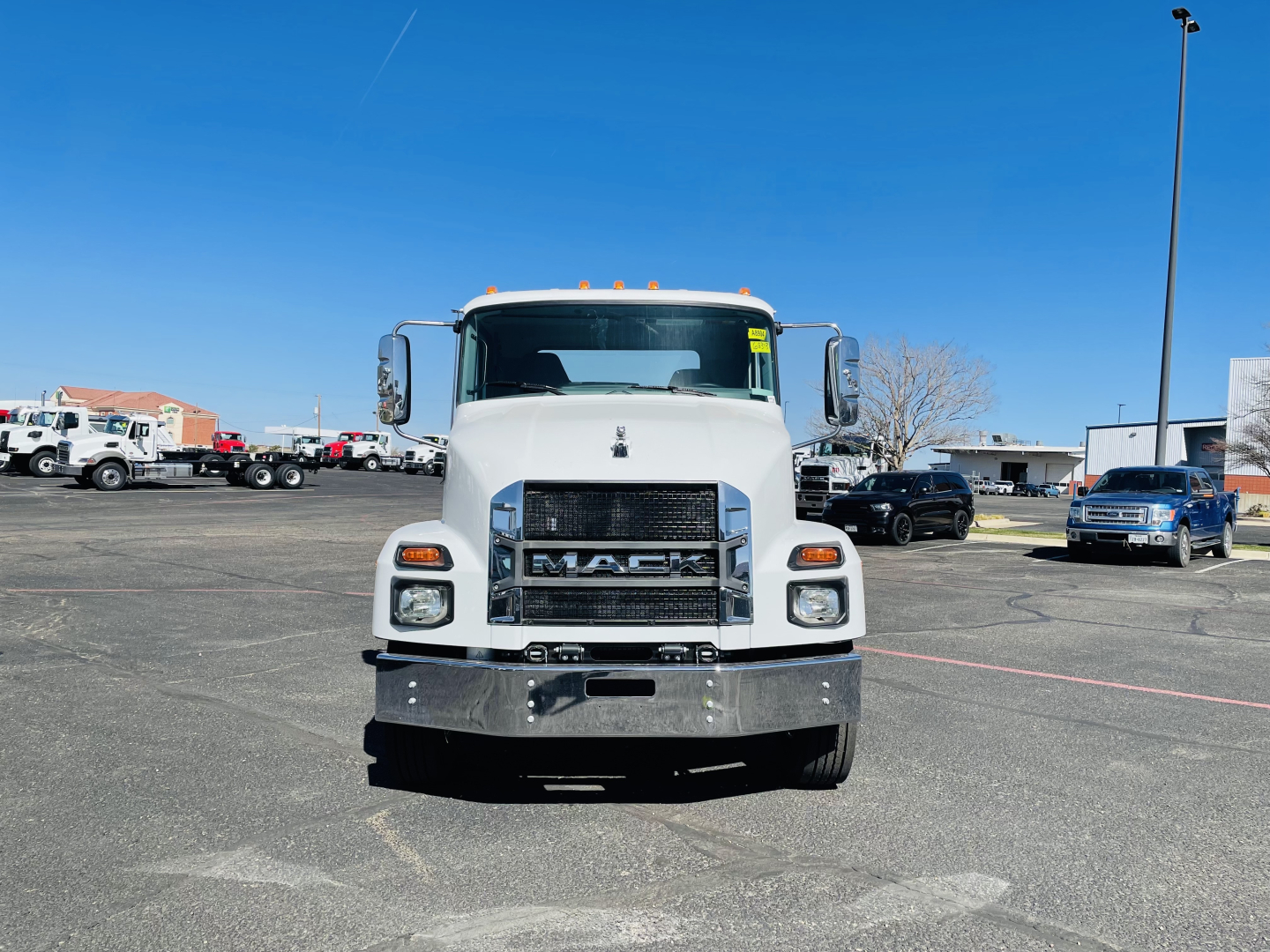 Front view of a white 2026 Mack MD6 flatbed truck showing chrome bumper, large grille with MACK emblem, dual headlights, and side mirrors.