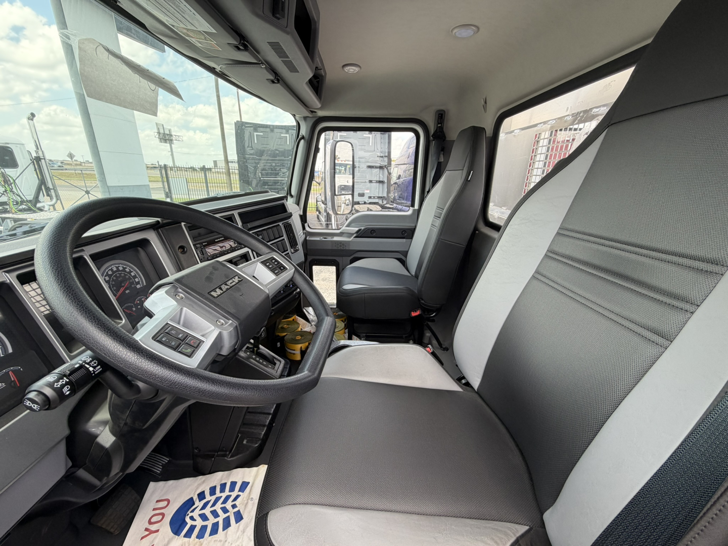CMV cab interior showing steering wheel, dashboard, and front seats with gray fabric upholstery; driver seat area and windshield with part of exterior visible.
