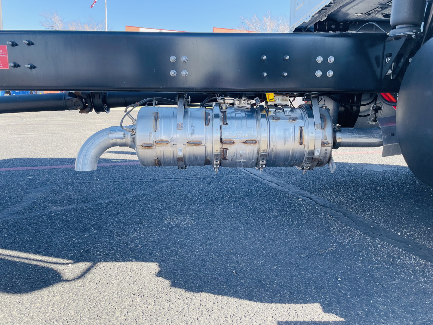 Underbody view of a Mack MD6 flatbed truck showing a cylindrical exhaust or fuel treatment component with pipes and mounting brackets.