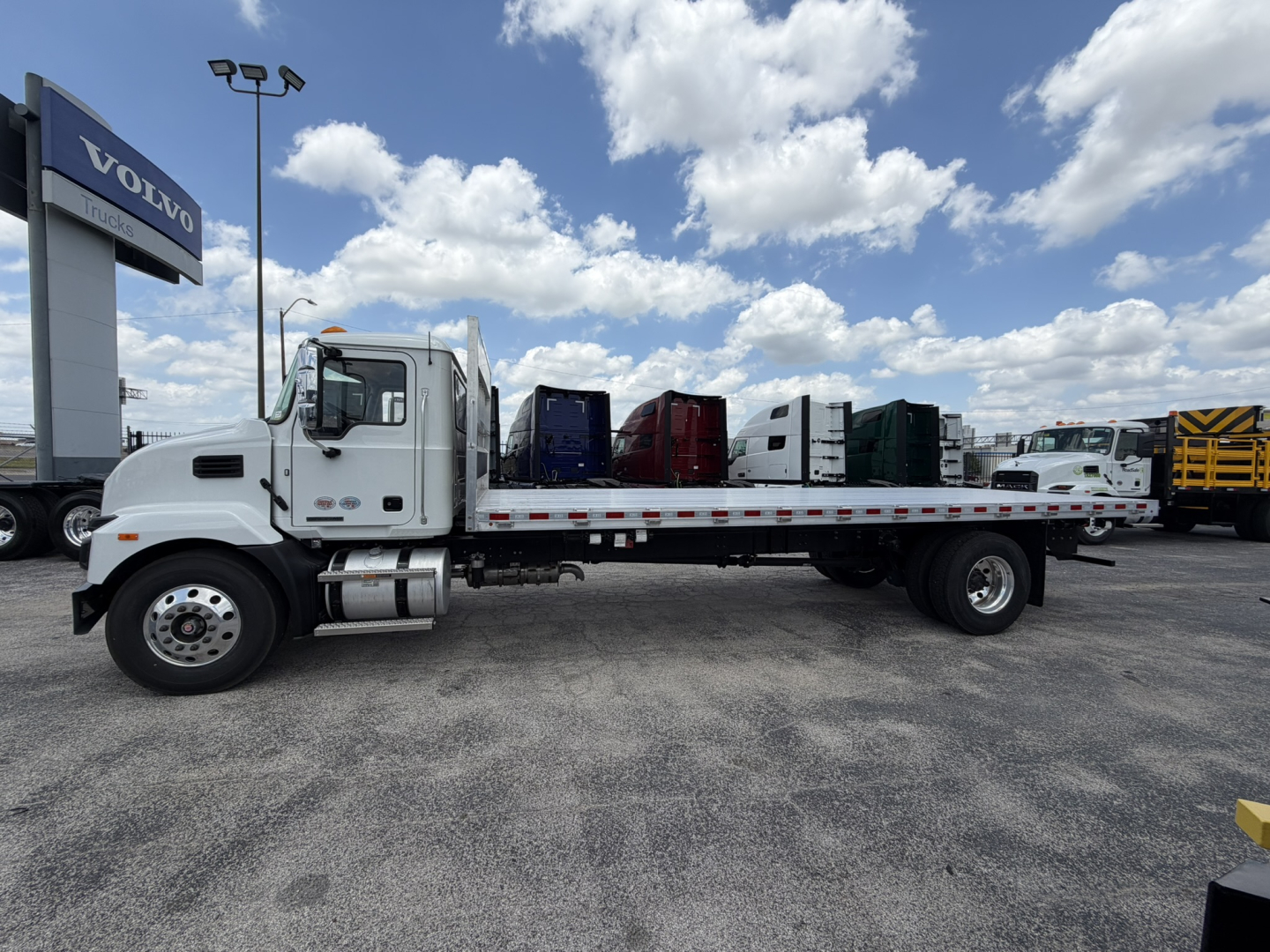 Flatbed truck side view showing a white 2026 Mack MD6 straight truck with a long flatbed, cab, dual rear axles, and a Volvo dealership sign in background.