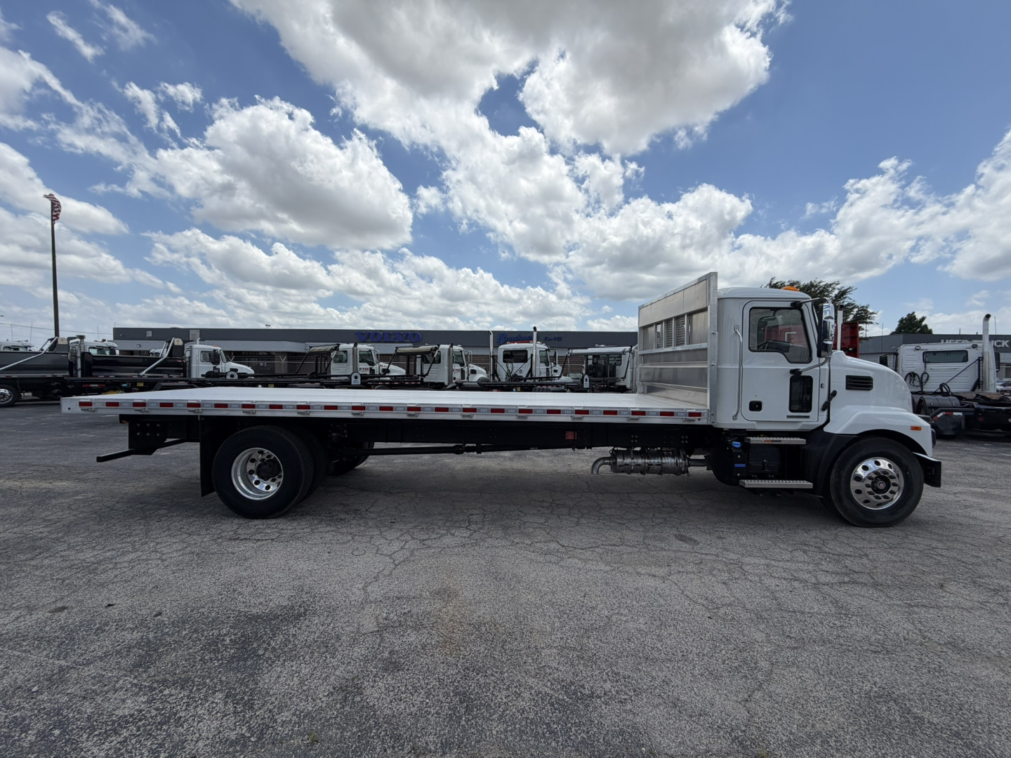 Flatbed truck side view showing white Mack MD6 cab and long flatbed with wheel, side rail, and cargo area.