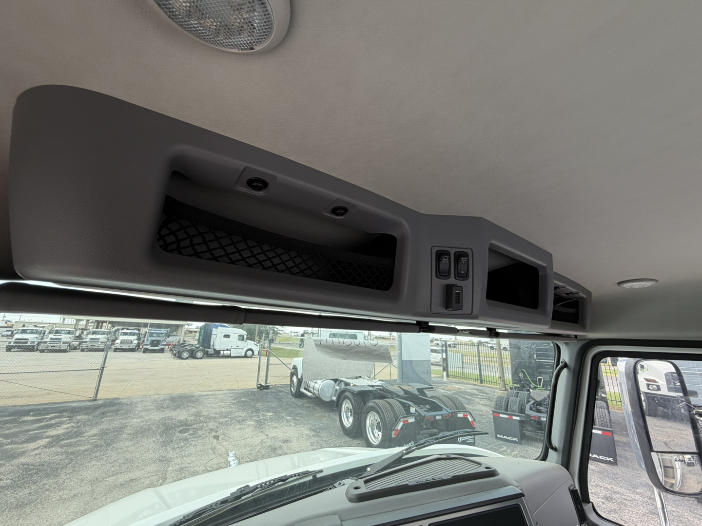 Cab interior showing overhead storage panel and control switches, windshield and side mirror, exterior lot with trucks in background.