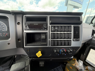 Dashboard of a 2026 Mack MD6 flatbed truck showing radio, climate controls, multiple switches, and yellow hood release handle.