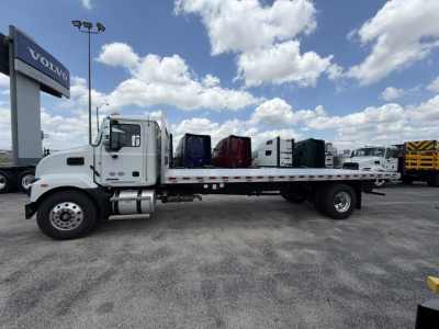 Flatbed truck side view showing a white 2026 Mack MD6 straight truck with a long flatbed, cab, dual rear axles, and a Volvo dealership sign in background.