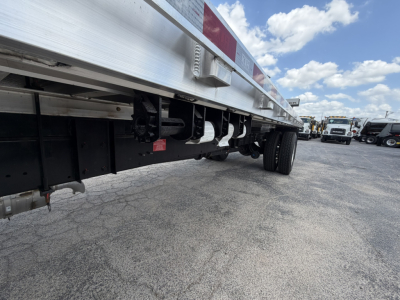 Flatbed truck undercarriage showing suspension, frame and side railing with red reflective strip. Multiple trucks in background.