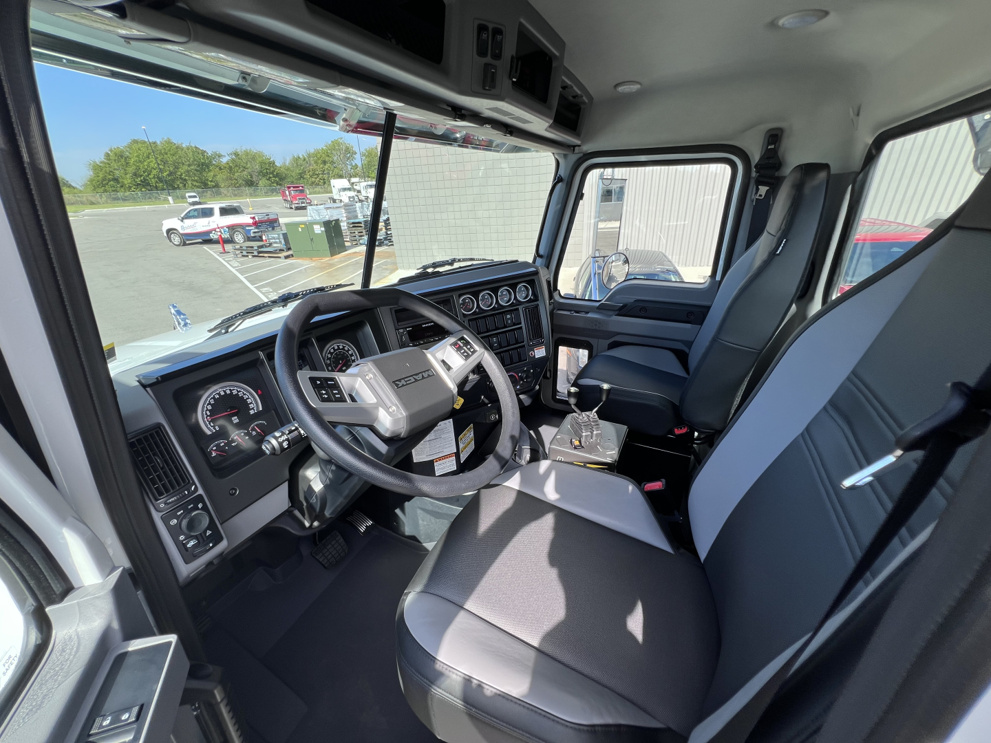 Truck cab interior of a 2026 Mack GR64B with steering wheel, dashboard gauges, and two-tone seats; view from the driver’s door; overhead compartments and window in the background.