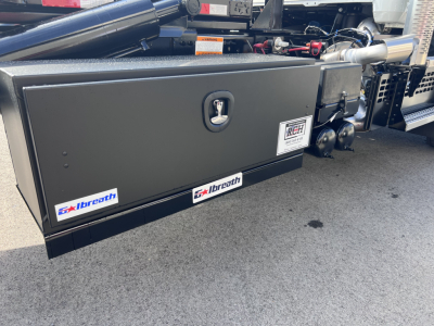 Grainy flatbed side toolbox on Mack GR64B rolloff truck showing toolbox, hydraulic lines, exhaust stack, and stickers