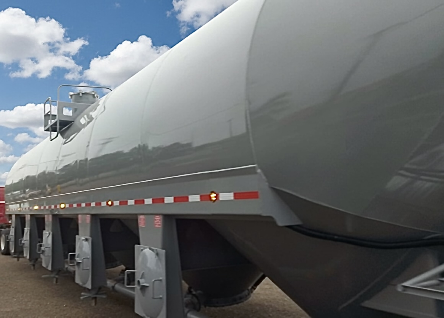 Pneumatic dry bulk trailer with large cylindrical cement storage silo on a multi-axle trailer, reflective gray exterior, safety ladders and red/white reflective stripes.