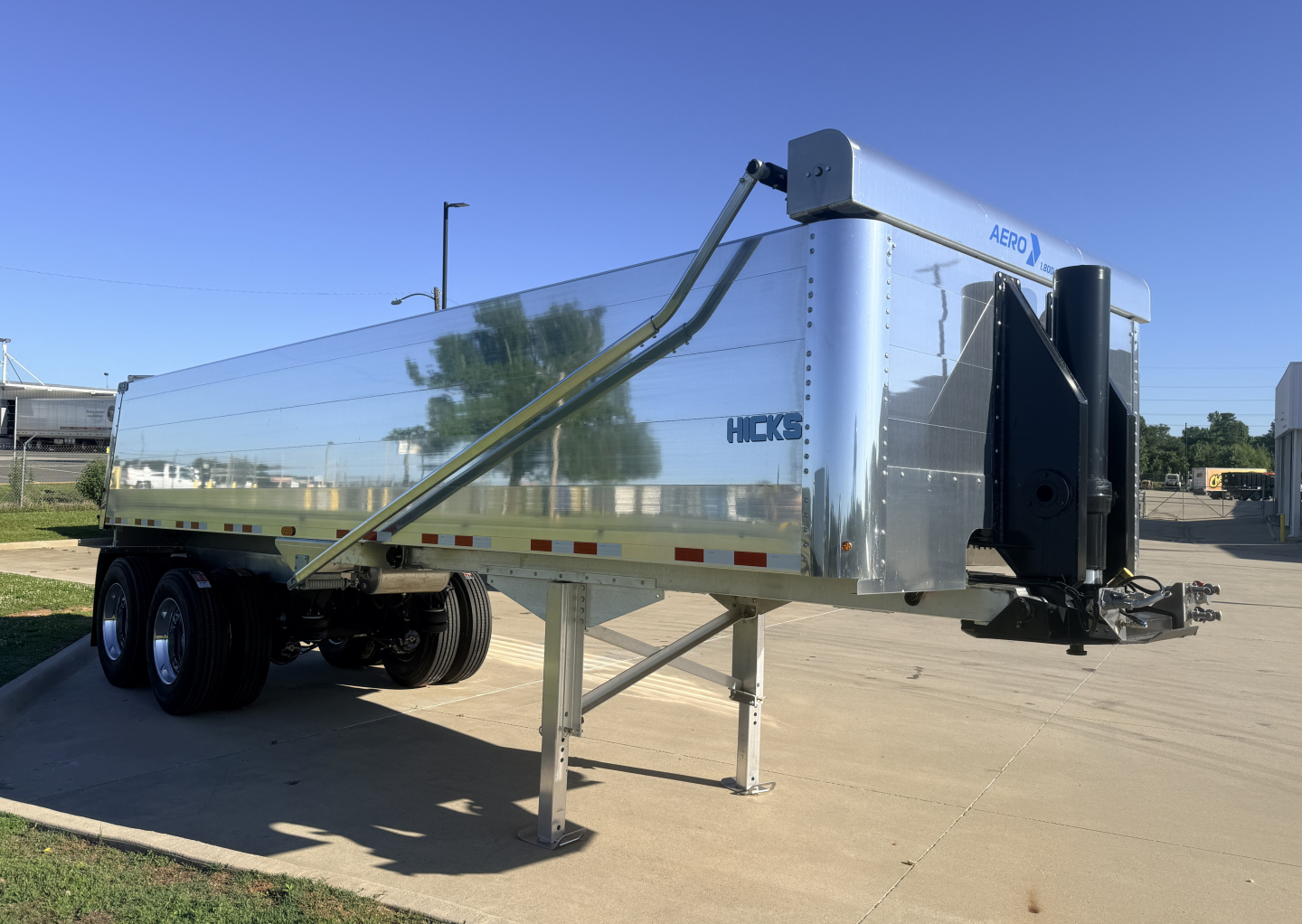 Hicks dump trailer with polished aluminum exterior, dual rear axles, and front hoist mechanism visible.