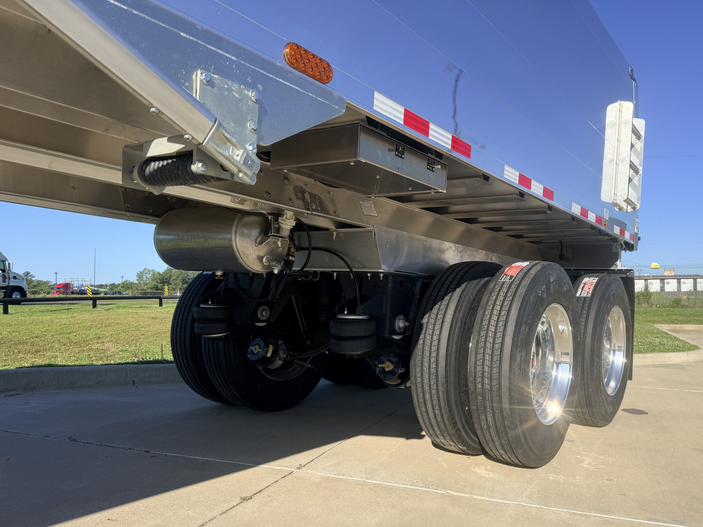 Dump trailer rear/side view showing dual rear axles with tires, undercarriage, suspension components, and a cylindrical tank or axle housing. No visible text readable aside from the trailer structure.