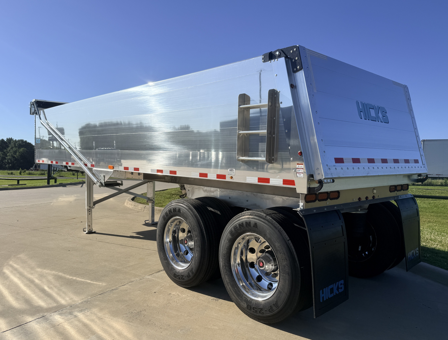 Hicks dump trailer with polished aluminum side, dual rear wheels, and ladder on front panel showing HICKS logo.