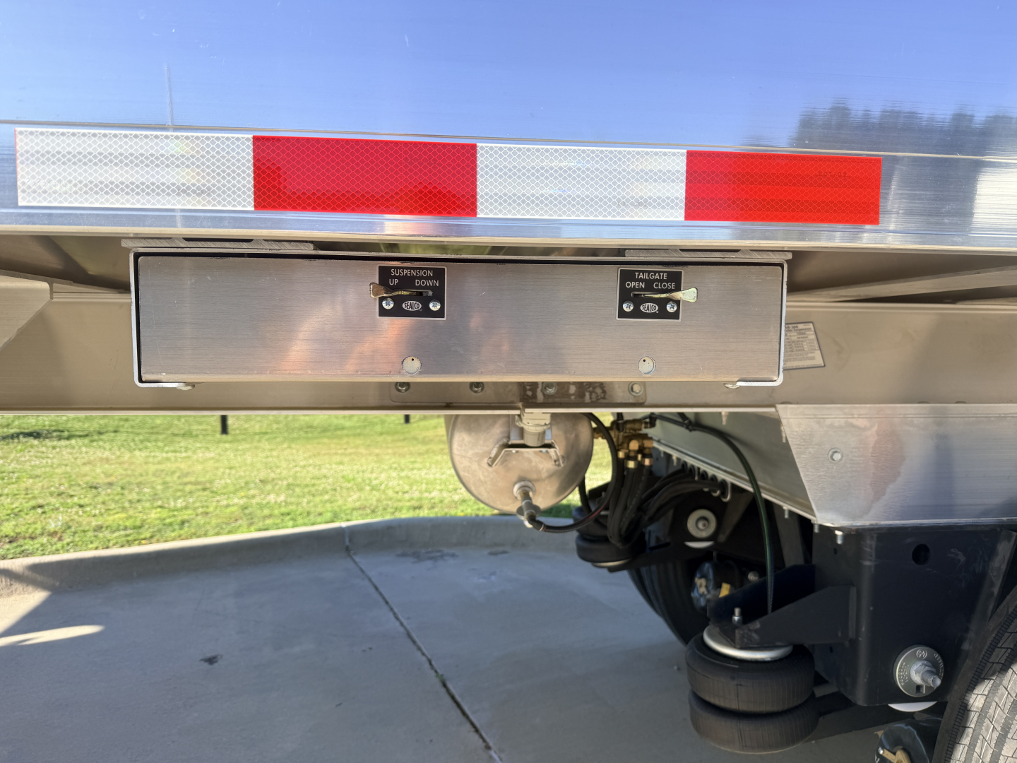 Trailer rear showing toolbox with two latches and labels, undercarriage with coiled hoses and a round hitch, reflective red/white safety tape along top edge.