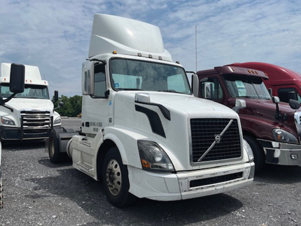 Volvo VNL 300 daycab tractor front view showing large grille, headlights, white paint, and other trucks in background.