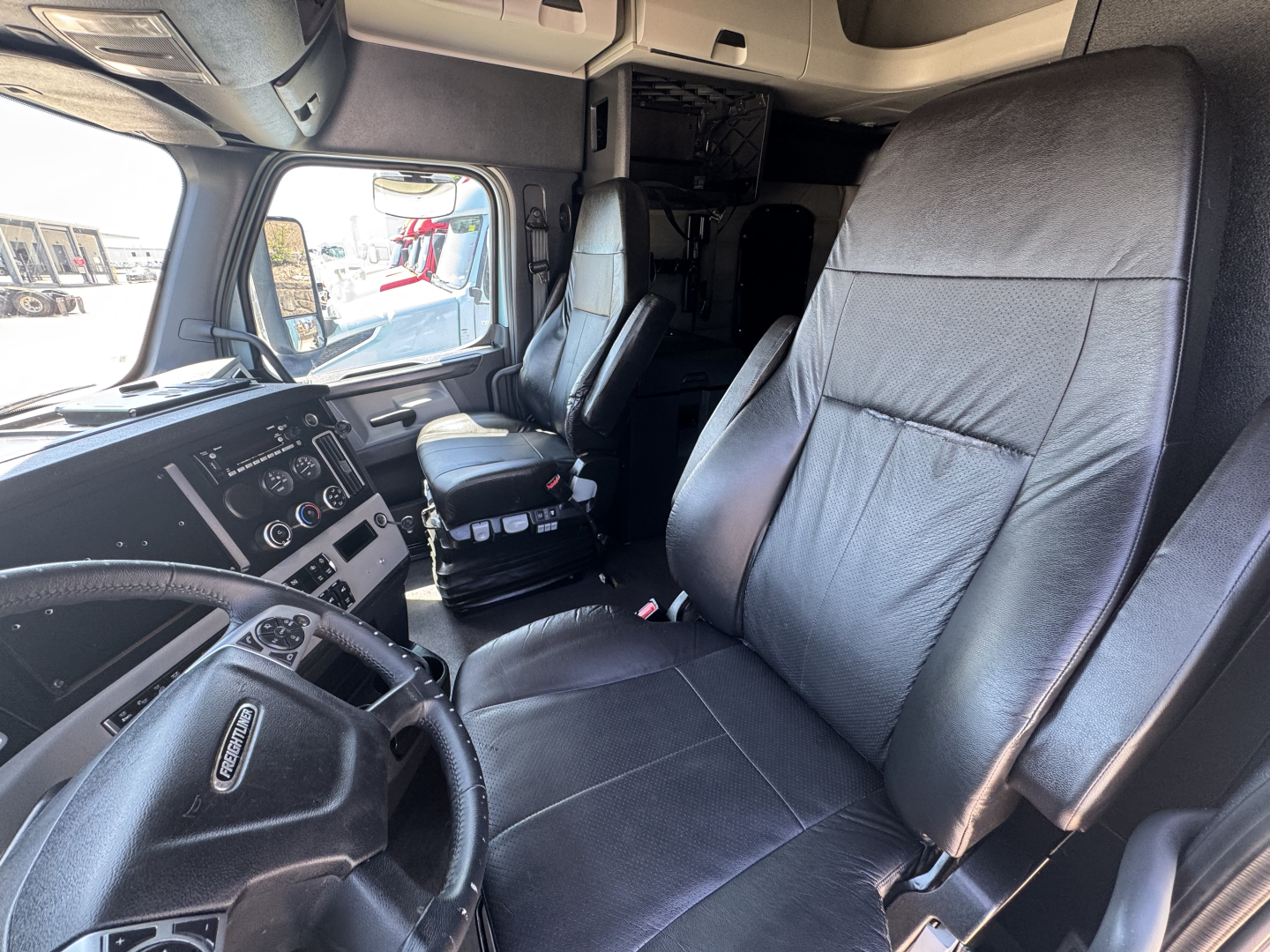 Cab interior of a 2023 Freightliner Cascadia 126 conventional sleeper showing driver seat, passenger seat, dashboard with steering wheel and control panel, and overhead compartments.