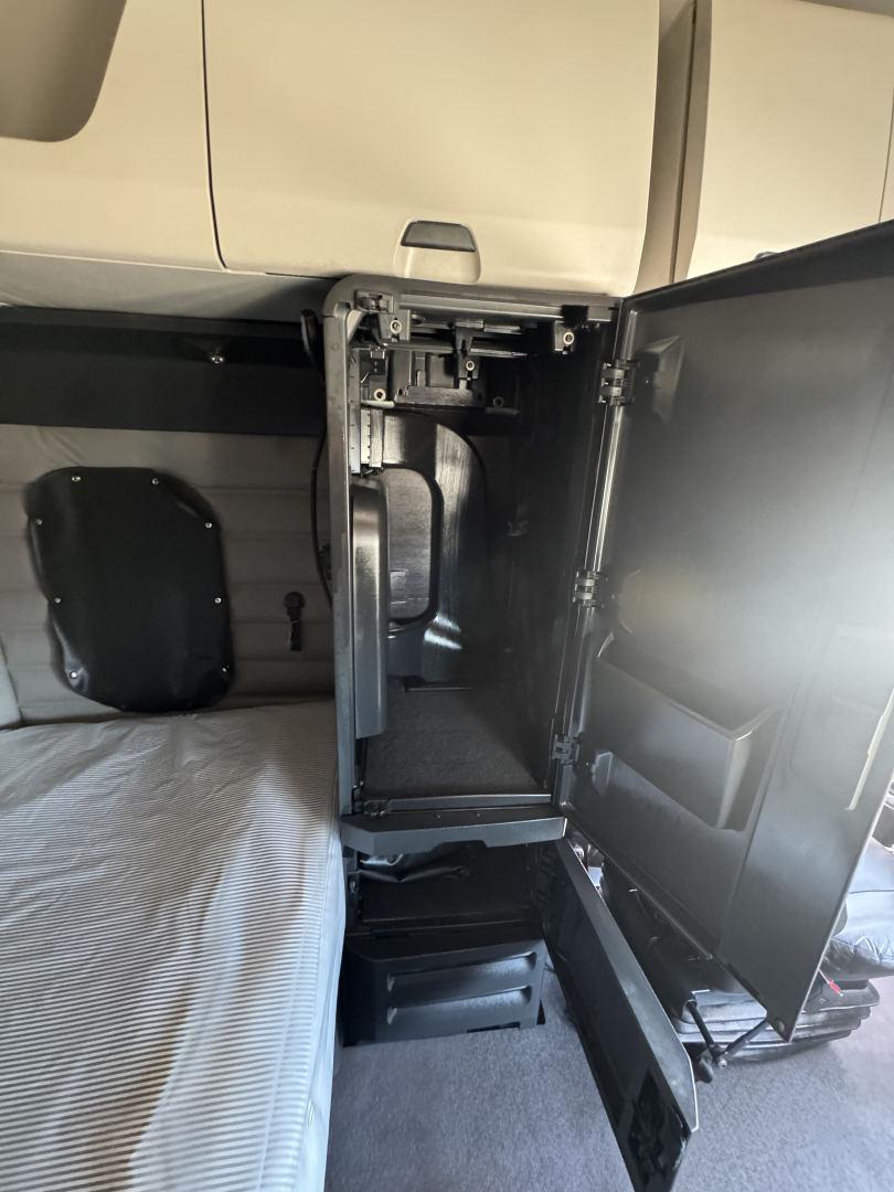Cab interior of a Freightliner Cascadia 126 showing a storage/drawer area with a backpack-style bed and a side cabinet open; includes metal hinges and exposed frame.