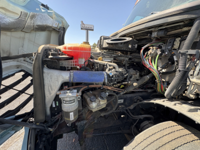 Truck engine bay of a 2023 Freightliner Cascadia 126 with exposed hoses, wiring, and fluid reservoirs visible from the left-front side.
