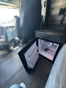 Freightliner Cascadia 126 cab interior showing open compact under-cab refrigerator with small freezer and two-door mini-fridge and door shelves. View includes driver seat and dashboard area.