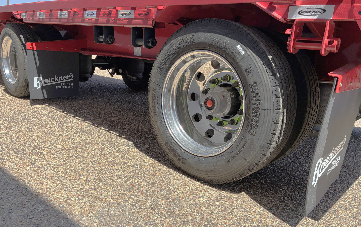 Trailer wheel and mudflap area of a red Dura Haul drop deck trailer with chrome wheel, black mudflaps, and branding stickers.