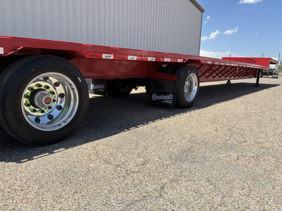 Trailer side view showing red drop deck trailer with Brackner aluminum wheels and reflective stickers, black tires, gravel ground, storage building in background.