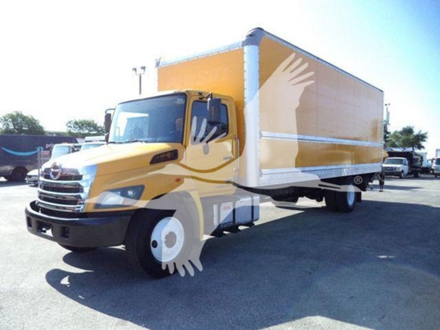 Box truck with yellow cab and large orange box, viewed from front-left, showroom lot in background.
