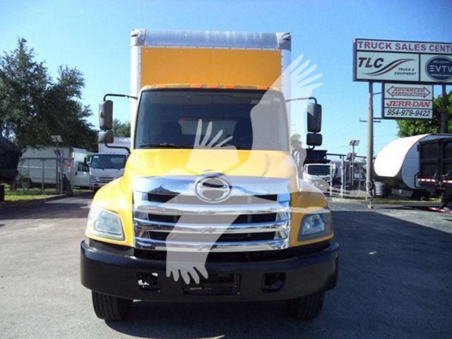 Front view of a yellow Hino 268A box truck. Hood emblem, chrome grille, headlights visible; side mirrors; cargo box in yellow; dealership signs in background.