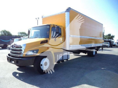 Box truck with yellow cab and large orange box, viewed from front-left, showroom lot in background.