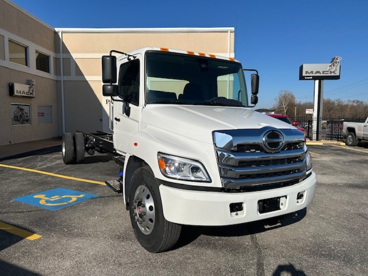 Truck cab and chassis showing front view of a white 2025 Hino L6 with chrome grille and Hino emblem, daytime outdoor lot.