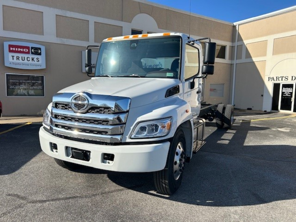  cab and chassis truck showing front grille and Hino emblem, white exterior, parked outside dealership with sign in background.