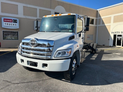  cab and chassis truck showing front grille and Hino emblem, white exterior, parked outside dealership with sign in background.