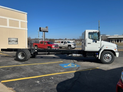Truck cab chassis showing white Hino truck with long chassis frame and exposed drivetrain, side view, dealership lot in background.