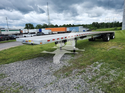 Flatbed trailer from Reitnouer Big Bubba line, low ground clearance with dual rear axles, aluminum decking, reflective safety tape along sides, set on grassy lot.