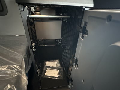 Cab interior showing open storage compartment with shelves inside a new Mack PR64T76H truck cab, black interior, plastic-wrapped seat nearby.