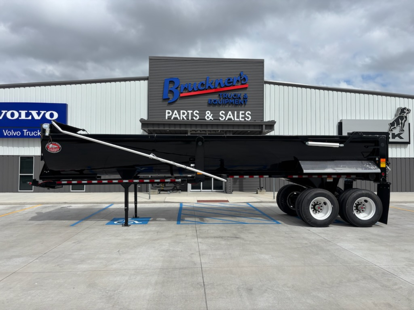 Dump trailer with black body and raised side rail, three-axle configuration, showing Bracken's Parts & Sales building in background.