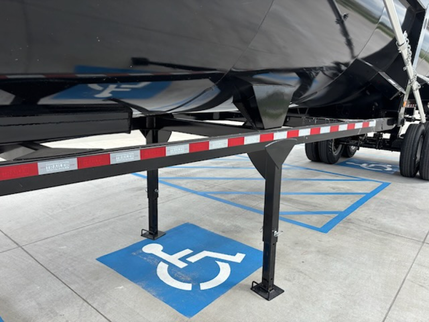 Trailer side view showing a black dump trailer with support legs and a blue wheelchair-accessible parking space symbol on the ground.