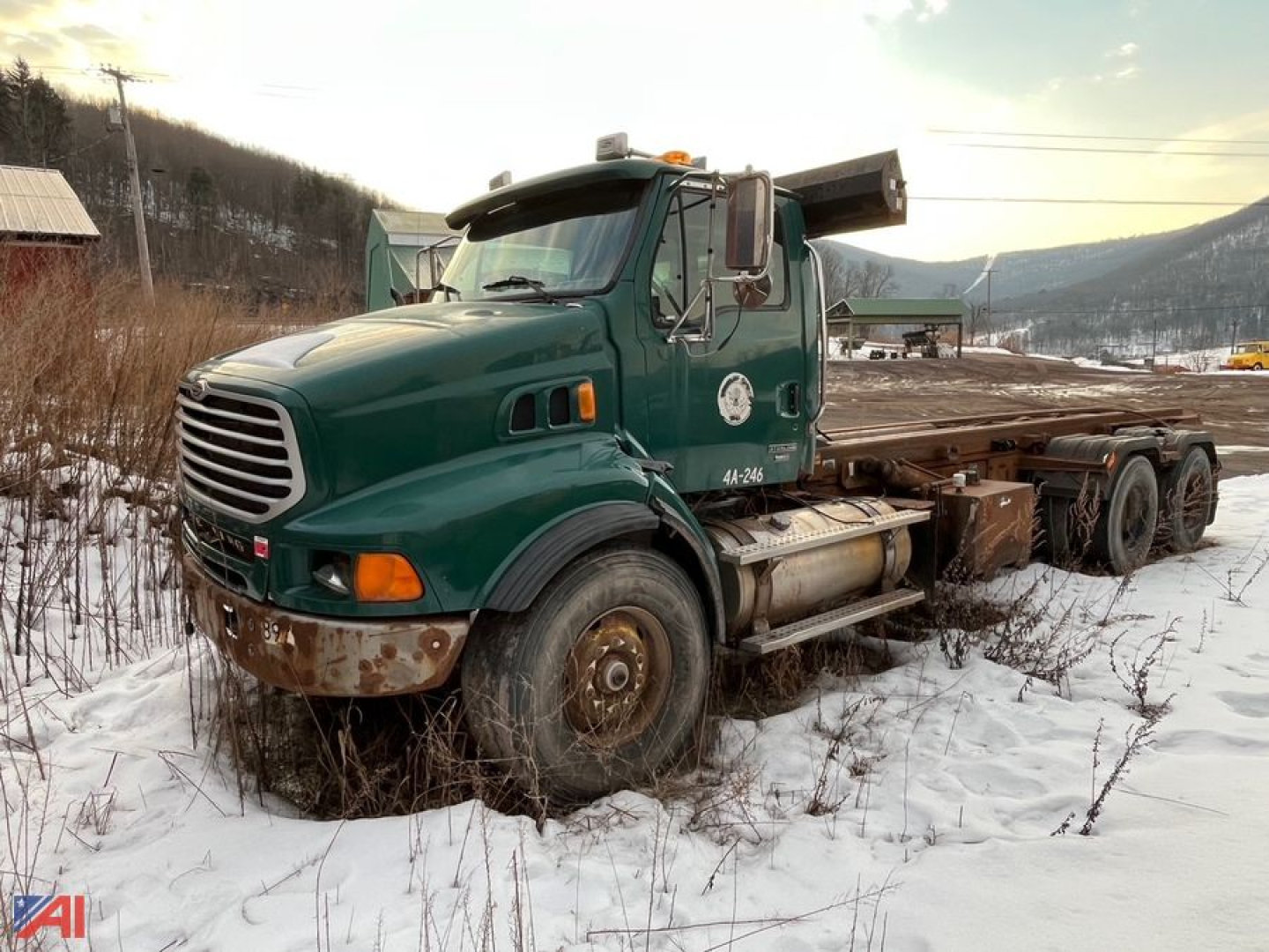 Green Sterling LT9500 roll-off truck in a snowy lot, front grille, cab with logo, with a flatbed and wheel chocks; visible heavy-duty tires and rust on bumper.