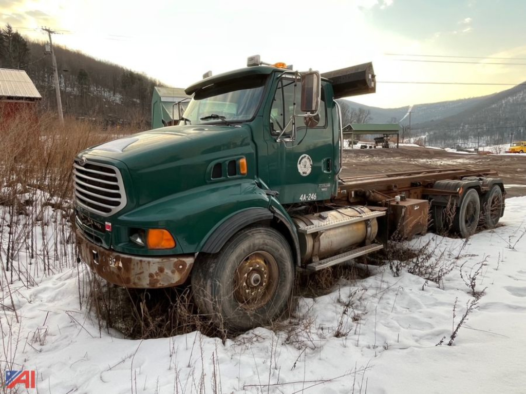 Green Sterling LT9500 roll-off truck in a snowy lot, front grille, cab with logo, with a flatbed and wheel chocks; visible heavy-duty tires and rust on bumper.
