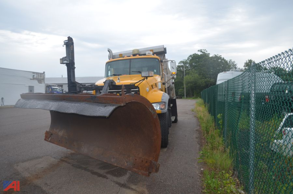 Electric yellow Mack Granite dump truck with a large front plow, outdoor lot, chain-link fence on right.